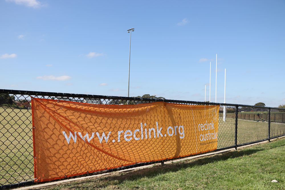A football field on a sunny day, with goal posts to the right. An orange banner on the fence reads www.reclink.org and has the Reclink logo