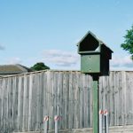 A green-painted timber letterbox in front of a domestic fence. There's a blue sky behind and some green trees.